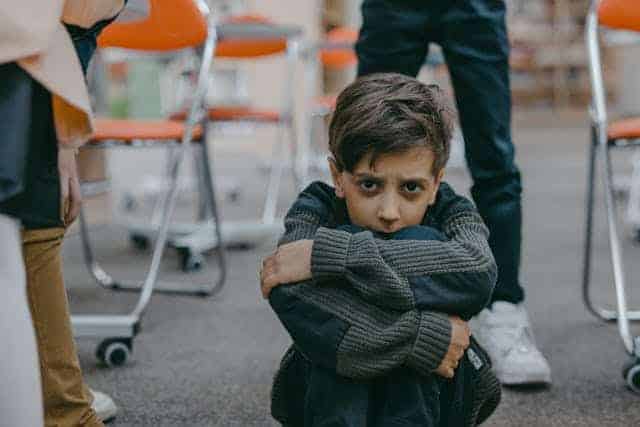 Scared-boy Mass deportation boy wearing a gray sweater sits on the ground, arms around his knees, looking scared.